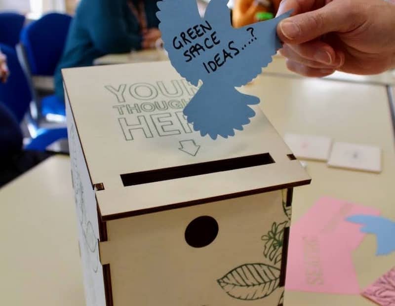 Hand holding bird-shaped note with 'green spaces ideas' on it and bird box